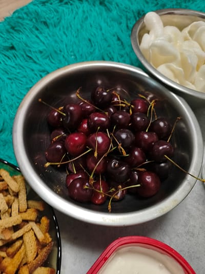 A high-angle shot shows a collection of food items arranged on a surface, likely a table. In the foreground, a stainless steel bowl overflows with dark red cherries. The cherries are glossy and have long, thin stems. To the left of the cherry bowl, a dark bowl filled with toasted bread pieces, appearing to be croutons or rusks, is partially visible. In the background, to the right, another stainless steel bowl contains a pile of white, puffy, food items, possibly puffed rice or a similar snack. A bright teal, fuzzy textile is visible in the upper left corner of the image, providing a contrasting texture and color. The lighting suggests it is daytime, with no specific weather conditions or time of day discernible. The setting appears to be indoors, given the artificial lighting and the nature of the food items. There is no visible text in the image. The overall impression is a presentation of ingredients or snacks, possibly for a meal or gathering.