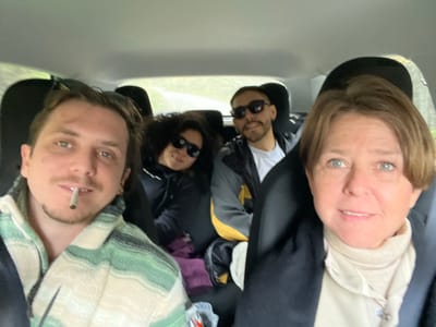 The image depicts five people in the interior of a vehicle. The perspective suggests the photograph was taken from above. A fair-skinned, mature woman with short brown hair is on the left. Next to her is a man with sunglasses, wearing a white t-shirt. Beside them is a woman with dark curly hair and sunglasses. To the right is an assortment of bags and clothes. On the right is a man with short facial hair, wearing a white and green patterned shirt and smoking a rolled cigarette. The vehicle's interior is primarily gray. The location, Puerto Almanza, Argentina, cannot be confirmed through the imagery alone.