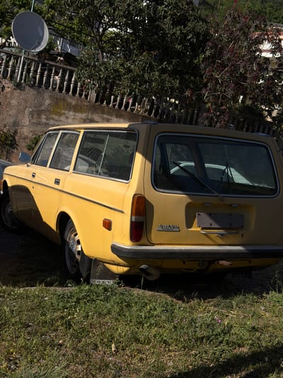 A yellow Volvo station wagon is parked on an overgrown roadside in Stupovi, Montenegro. The vehicle is positioned adjacent to a stone wall and a variety of vegetation, including grass and a tree with dark foliage. A satellite dish is visible in the background, attached to a building. The scene appears to be taken in daylight.
