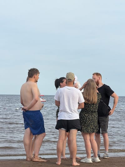 A group of four young adults are standing on a sandy shore next to a body of water. The water appears to be a lake or the sea, with ripples indicating a light breeze. The sky is overcast, suggesting it is either early morning or late afternoon, or simply a cloudy day. One man in the foreground is shirtless, wearing blue swim trunks. He is standing with his back to the camera, looking out at the water. Behind him, another man in a white t-shirt and black shorts is also facing away, with a woman in a floral dress and a man in a black t-shirt and distressed denim shorts standing to his left. The woman in the floral dress is facing the camera, her hair obscuring her face. The man in the black t-shirt is looking down at her. Another person, partially obscured, is between the man in the white t-shirt and the woman in the floral dress. The group appears to be a mix of couples or friends enjoying a day by the water. There are no visible signs or text in the image. The overall mood seems relaxed and casual.