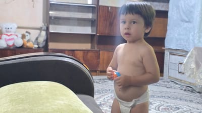 A young child, wearing a diaper, stands in a room. The child has dark hair and is looking to the left with a curious expression. The child is holding a small toy in their hands. The room appears to be a living area, with a couch visible in the foreground and wooden furniture and shelves in the background. Several stuffed animals are on a shelf in the background. The lighting suggests it could be daytime, with light coming from an unseen source. The overall impression is a casual, indoor scene of a child playing at home. The location context provided is Yakkabog, Uzbekistan.