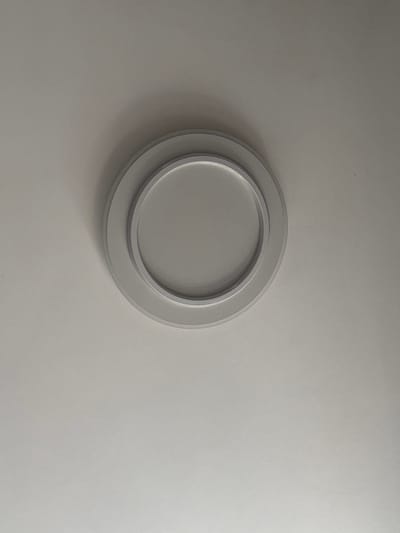 The image shows a close-up, overhead view of a recessed ceiling light fixture. The fixture is circular and has a layered design with a matte white outer ring and a slightly recessed, lighter gray inner circle. The surrounding ceiling is a uniform, light gray color, with subtle shadows cast by the fixture, indicating the light source is overhead. There is no indication of the location being in Vsevolodovo, Russia, or any other specific city. The scene is static and devoid of any people or actions.