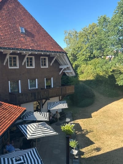 An aerial, high-angle shot captures a view of a multi-story building with a red tiled roof, possibly a guesthouse or hotel, in St. Märgen, Germany. The building has dark brown wooden siding and a balcony with a wooden railing. Several outdoor seating areas are visible on the ground floor, furnished with black metal chairs and tables, some shaded by black and white striped umbrellas. Two people are seated at one table under an umbrella, while another person is seen relaxing in a chair on a lower patio. A pathway winds through a dry, sunlit grassy area adjacent to the building, bordered by dense green trees. The sky is clear and blue, suggesting a sunny day.