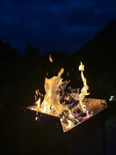 An outdoor fire pit burns brightly at night, with flames licking upwards from burning logs and embers. The fire pit is made of dark metal and sits in what appears to be a garden or yard, with dark trees and foliage visible in the background. In the distance, the silhouette of a mountain range is visible against a dark blue, cloudy sky. The scene suggests a peaceful evening, possibly in a rural or suburban area. The location is specified as the municipal unit of Kassopaia, Greece.