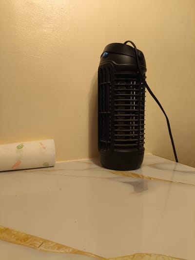 The image shows a black, cylindrical electric insect killer placed on a white, marble-patterned surface. The insect killer has a grille on its sides and a blue light near the top. A black cord extends from the top of the device and goes towards the right edge of the frame, disappearing out of view. To the left of the insect killer, there is a roll of white paper with a pattern of small orange carrots and green leaves printed on it. The background is a plain, textured wall painted in a light yellow or cream color. The lighting suggests an indoor setting, likely at night or in a room with artificial lighting. The scene is still, with no activity taking place. The overall impression is one of domesticity, with the insect killer and paper roll suggesting a functional rather than decorative purpose. The location context provided (Imus, Philippines) does not offer any specific visual cues within the image itself to confirm or deny this location. There is no visible text in the image.