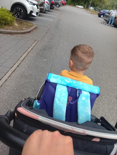 This is a first-person perspective photo taken in a parking lot, likely in Chemnitz, Germany. The foreground shows a hand gripping the handle of a stroller, with the back of a young child visible in the stroller seat. The child is wearing a yellow t-shirt and has short brown hair. Attached to the back of the stroller is a blue backpack with light blue straps adorned with white snowflake-like patterns. The stroller itself is predominantly black with some pink and white accents on the fabric.
The background reveals a paved parking lot with several cars parked in designated spots. To the left, the front of a white SUV is visible, parked next to a small grassy area with a concrete border. Further down the parking lot, other vehicles, including a dark blue car, are present. In the distance, trees and foliage suggest an outdoor setting. The lighting indicates it is daytime, and the overall scene appears to be a typical day in a public parking area, possibly near a shopping center or park. No text is clearly visible in the image.