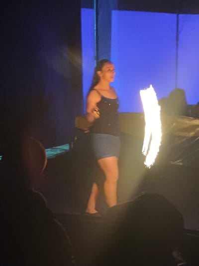 A woman in a black tank top and denim skirt stands on a stage with a blue background. She is facing slightly to the right, looking off into the distance. There is a bright, elongated flame or light source beside her. The foreground is dark, with the silhouettes of heads presumably belonging to audience members. The lighting casts strong shadows and highlights, obscuring some details of the scene.