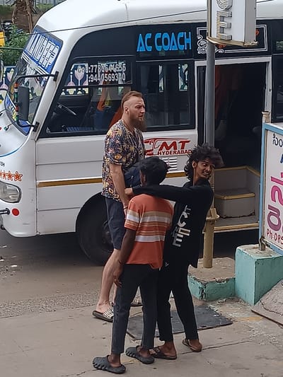 This media file captures a street scene in Kurnool, India. Three individuals are the primary subjects. On the left, a man with a beard and a distinctive haircut stands wearing a colorful patterned shirt and dark shorts. To his right, two younger men are interacting. One, in an orange and white striped shirt and dark pants, is partially obscured. The other, wearing a black long-sleeved shirt with white lettering down the side and dark pants, has his arm around the first young man and is looking back at the camera with a smile.
They are standing on a paved surface next to a white bus that appears to be an AC coach. The bus has "AC COACH" and "HYDERABAD" written on its side, along with phone numbers and other text, some of which is visible in the OCR. The bus has a door open, revealing steps leading inside. A sign with some script and "Ph:085" is visible on the right.
The scene appears to be during the daytime, with natural light suggesting it's not particularly early or late. The overall atmosphere seems casual, with the young men interacting playfully. The setting is urban, with the bus, pavement, and the bus stop structure indicating a public transportation area.