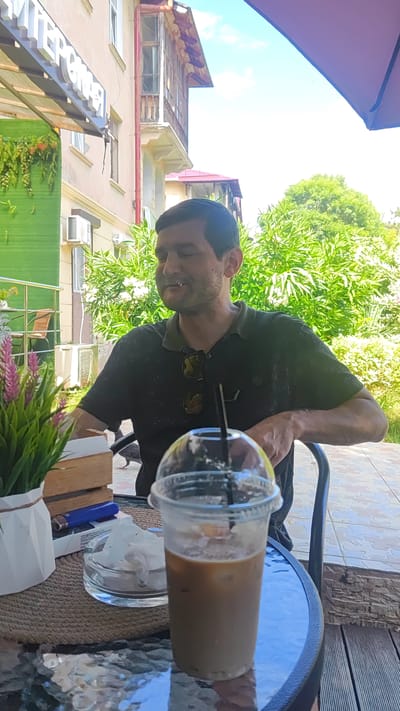 A man is sitting at a table outside, possibly at a cafe in Dagomys, Russia, based on the context provided. He is wearing a dark green polo shirt and has sunglasses clipped to the collar. In front of him is a plastic cup with an iced coffee and a straw. To his left, on the table, is an ashtray with a cigarette, a pack of cigarettes, and a lighter. A potted plant with pink flowers is also on the table, to the left of the coffee. Behind the man, there is greenery, a building with balconies, and a blue sky. The man appears to be in the process of smoking.