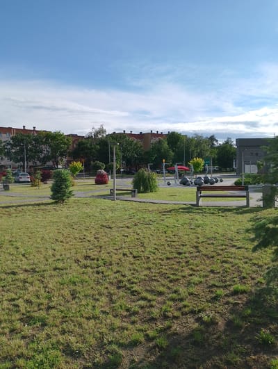 This is a daytime, eye-level, full-shot photograph of a park area in Kolno, Poland. The foreground is dominated by a grassy lawn with patches of dry, matted grass and sparse green growth. In the mid-ground, a paved pathway winds through the park. To the right of the pathway sits a wooden bench with a dark brown seat and grey concrete supports. Further into the mid-ground, a playground structure with red metal supports and grey spherical objects is visible. Several large, dark grey, spherical objects are arranged on the ground near the playground. To the left of the pathway, a small evergreen tree stands, and beyond it, a variety of bushes and trees are planted, including a vibrant red and pink floral display. In the background, a multi-story brick apartment building with a red-tiled roof and visible chimneys is situated behind a line of trees. Another modern building with dark cladding and a pitched roof is visible to the far right. The sky is a clear blue with scattered wispy white clouds. A silver car is parked on the road to the left of the park.