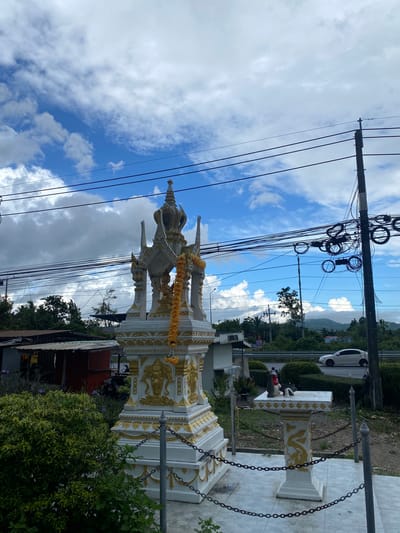 The image depicts a spirit house decorated with garlands and elephant carvings, located in Ban Ko Pru, Thailand. Overhead, there are power lines against a partly cloudy sky. A road with a passing white car runs adjacent. There are bushes surrounding the spirit house and part of a corrugated iron roof is visible behind it. Another shrine can be seen in the background.