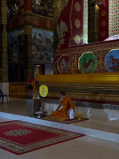 A Buddhist monk in orange robes sits on a mat on the floor of a temple in Na Nuea, Thailand. He is facing to his left and appears to be performing a ritual. In front of him are various religious items, including a small golden bowl and a piece of paper. Behind the monk, a large golden drapery hangs across the temple wall, adorned with intricate patterns. The temple walls are decorated with colorful murals depicting scenes from Buddhist stories and motifs. A red carpet with a traditional pattern is laid out on the floor in front of the seating area. In the background, another person in a red shirt and dark shorts walks away from the camera. The lighting suggests it is daytime, with sunlight filtering into the temple, creating shadows on the tiled floor.