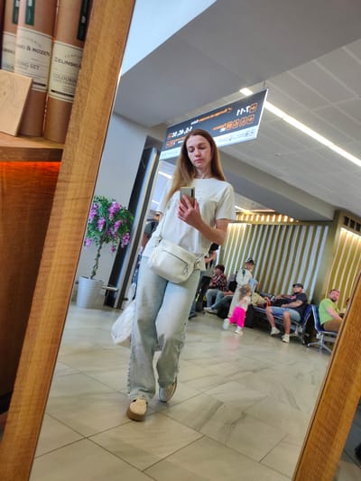 The image is a reflection in a large, wooden-framed mirror, showing a woman in a white t-shirt and light blue wide-leg jeans holding a smartphone and a white purse. She is standing indoors, likely in an airport, as suggested by the signage overhead that appears to be in Cyrillic and includes directional arrows and numbers, consistent with airport gate information. In the background, other people are visible sitting on benches, waiting. A child in bright pink pants is running in the foreground of the reflected scene. The environment appears to be a modern indoor space with tiled flooring and striped wall paneling. The mirror itself is positioned to capture the woman, the reflection of the waiting area, and a shelf to the left with cylindrical items that resemble product packaging.