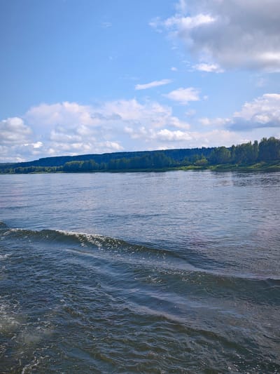 The image depicts a wide river with gentle waves in the foreground, suggesting movement, possibly from a boat. The water is a deep blue-grey, reflecting the sky. In the midground, the opposite bank of the river is lined with a dense forest of green trees, with a faint shadow cast by a distant hill or mountain range on the horizon. The sky above is a vibrant blue, dotted with fluffy white and grey clouds, indicating fair weather. The scene is serene and natural, likely capturing a moment of travel or observation from the river. The location is identified as Kargino, Russia.