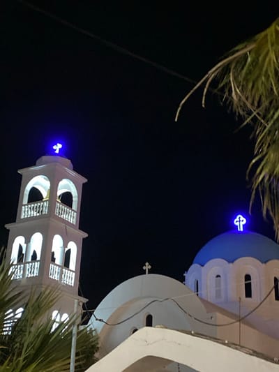 The image displays a white Greek Orthodox church at night in Megalochori, Greece. The scene is dominated by two main structures: a blue-domed section on the right and a white bell tower on the left. The blue dome is topped with a brightly illuminated blue cross. The white bell tower features multiple arched openings with internal white illumination and is also crowned by a brightly illuminated blue cross. A third, unlit white cross is situated atop a smaller white dome positioned between the bell tower and the main blue dome. The architectural elements are predominantly white, sharply contrasting with the deep black night sky. Green palm fronds partially obscure the lower left foreground and extend into the upper right portion of the frame. Thin, dark utility lines are visible horizontally across the dark sky, above the church structures. No individuals are present in the image.
