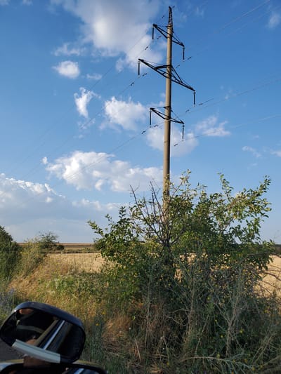 The image depicts an outdoor scene under a bright blue sky with scattered white clouds. A tall, concrete power line pole stands prominently in the center, supporting multiple electrical wires that extend across the frame. In the foreground, on the right, is a dense bush with green leaves and small yellow flowers. To the left, a car's side mirror is visible, reflecting a person and the surrounding environment. Beyond the vegetation and the power pole, a flat, golden field stretches towards the horizon, suggesting a rural or agricultural area. The overall impression is one of a clear, sunny day in a semi-rural setting, possibly on the outskirts of a town like Konstantinovka, Moldova.