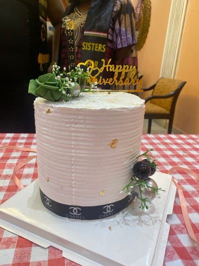 The image shows a white cake with a gold "Happy Anniversary" topper, a black Chanel ribbon around the bottom, and a white and green floral decoration. The cake is on a white cake stand on top of a red and white checkered tablecloth. It is likely located in Buea, Cameroon, based on the provided coordinates. The coordinates correspond to an area with a mix of residential and commercial areas in Buea, known for its vibrant cultural scene.