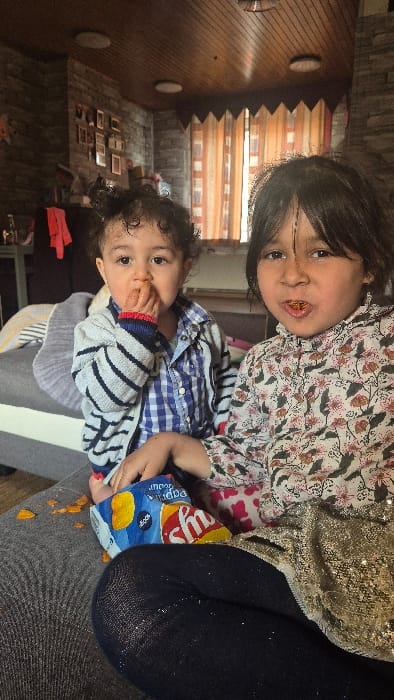 This image depicts the interior of a residence in Melle, Belgium. Two children are seated on a grey upholstered sofa. The younger child, positioned on the left, wears a blue and white checkered shirt beneath a grey and white striped cardigan, with their right hand at their mouth. The older child, on the right, wears a light-colored floral patterned shirt and dark, possibly metallic-threaded, leggings, with orange food particles visible in their mouth. A blue bag of "Lay's Paprika Flavour" potato chips rests on the sofa between the children, surrounded by scattered orange potato chip fragments. Partially visible on the older child's lap is a textured, scaled reptile, possibly a bearded dragon. The background features a wall with a stone-like texture, a wooden ceiling equipped with two recessed light fixtures, and a window draped with orange and white vertical striped curtains. Multiple framed pictures are affixed to the wall on the left.