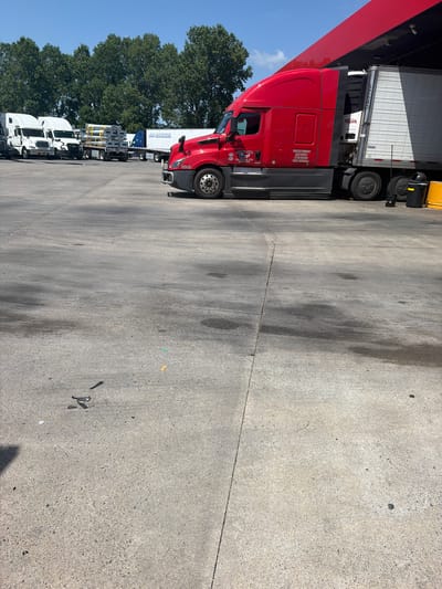 The image captures an outdoor scene in Hayti, United States of America, likely a truck stop or industrial yard, on a sunny day under a clear blue sky. In the foreground, a concrete surface shows signs of wear and tear with oil stains and scattered debris, including what appears to be nail clippers, a bottle cap, and small pieces of colored plastic.
Dominating the mid-ground is a large, red semi-truck parked next to a red overhang structure, possibly a canopy at a fueling station. The truck has the number "2649" visible on its side, and branding that includes "HOMELAND" and "TRAILER". A white refrigerated trailer is attached to the truck, displaying the number "52540" and "CALVIL". Two dark-colored trash receptacles, one black and one yellow, are situated near the rear of the truck.
In the background, several other white semi-trucks are parked, some with trailers. One trailer displays the text "J.R. SCHUGEL". A line of mature, leafy green trees forms a backdrop behind the trucks. The overall impression is one of a functional service area for commercial vehicles.