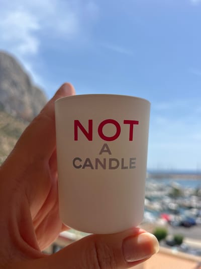 This media file is a close-up, eye-level shot of a person's hand holding a white, cylindrical object against a bright blue sky. The object appears to be a container for a candle, or possibly a candle itself. The text "NOT A CANDLE" is printed vertically on the object in bold red and grey letters. The "NOT A" is in red, and "CANDLE" is in grey. The person's hand is holding the object from below, with the thumb and fingers gently cupping its base. A fingernail, neatly manicured and with a light polish, is visible in the upper right corner of the frame.
The background is a vibrant, clear blue sky with a few wispy white clouds. In the distance, out of focus, there are hints of a coastal landscape with a body of water, possibly a marina with boats, and buildings or hills. The lighting suggests it is daytime, likely a sunny day, given the brightness of the sky and the shadows cast. The overall mood is casual and relaxed, possibly a vacation or leisure moment. The specific text "Calp, Spain" suggests the location of the photograph.