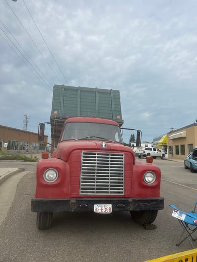This media file features a close-up, ground-level view of the front of a red truck. The truck has a large, chrome grille and a visible license plate that reads "82 B398 Alberta". The vehicle appears to be parked on an asphalt surface, possibly a street or parking lot, with a curb visible in the foreground. Behind the truck, several buildings are visible, suggesting an urban or commercial setting. One building has a yellow facade with windows and signage, and another is a lighter color with stone accents and windows. The sky is overcast with scattered clouds. There is a folding blue chair with a pink cup on it to the right of the truck. In the background, other vehicles, including a white pickup truck and a light blue car, are parked. The overall scene is daytime. The angle of the shot makes the truck appear dominant in the frame.