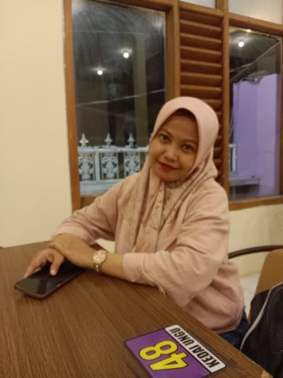 A single adult female is seated at a dark brown wooden table indoors, looking directly at the camera and smiling. She wears a light pink long-sleeved shirt, a matching light pink hijab, and a gold-toned watch on her left wrist. Her hands are clasped on the table. A black smartphone with a brown trim lies flat on the table to her left. To her right, a purple rectangular sign with yellow text "KEDAI UNGU" and the number "48" is displayed on the table. The background features a light cream-colored wall to the left and a window with wooden frames and wooden slat blinds to the right. Through the window, reflections of interior lights are visible, along with a white decorative railing and a distant purple-colored exterior structure. The scene is located in Pandean, Indonesia.