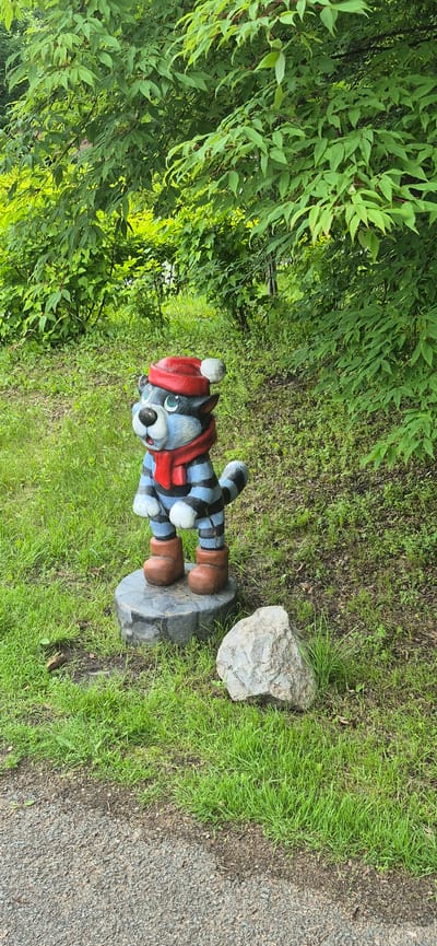 A whimsical, anthropomorphic cat statue stands on a grey, circular stone pedestal in a grassy area, positioned to the left of a large, grey rock. The cat is painted with blue and black stripes, wearing a red hat with a white pom-pom, a red scarf, and brown boots. It has a blue face with green eyes, a white muzzle, and a black nose. Behind the statue, lush green trees and foliage form a dense backdrop. The ground is covered in green grass, with patches of dark soil and leaf litter visible. A gravel pathway borders the lower left corner of the image. The overall scene appears to be an outdoor setting, possibly a park or garden, in Knevichi, Russia.