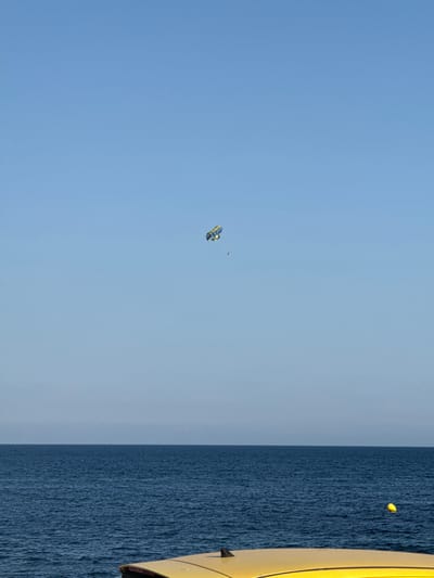 This is a vertically oriented, eye-level shot of a clear blue sky and the deep blue ocean. On the left side of the frame, the sky is a pale blue, gradually darkening towards the horizon. A small, yellow and blue parasail is visible in the upper portion of the sky, with a person attached to it. The parasail appears to be in motion, likely being towed by a boat.
On the right side of the frame, the ocean occupies the majority of the space, with a textured surface indicative of waves. A bright yellow object, possibly part of a boat or a buoy, is visible floating on the water's surface. The overall scene suggests a bright, clear day with good visibility, likely during daylight hours. The presence of the parasail and the ocean points to a recreational activity taking place at a coastal location. The cropped hints show the parasail in more detail and a closer view of the ocean and the yellow object. No visible text is present in the media. The location context of Aguadulce, Spain, suggests a warm, sunny climate suitable for such outdoor activities.