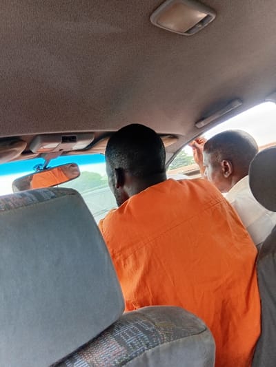 The image depicts the interior of a vehicle in Itobe, Nigeria, viewed from a rear passenger seat looking forward. In the immediate foreground, the back of a grey fabric car seat with a multi-colored patterned lower section is visible. Ahead, two individuals are seated. The primary subject is a person with dark hair, seen from behind, wearing a bright orange shirt or garment. To their right, a second individual is partially visible, with short, light-colored hair and wearing a white or light-colored shirt. This second person's right arm is raised, with the hand positioned near the upper part of the passenger-side window or door frame. The vehicle's interior features a light-brown textured headliner, an overhead light fixture, and a rearview mirror reflecting the orange shirt of the person in front. Through the blue-tinted windshield, green foliage and an open, bright outdoor environment are discernible, indicating daytime conditions. There are no overt interactions occurring between the individuals visible.