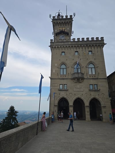The image shows the Palazzo Pubblico, a historic building in the city of San Marino, San Marino. The building is made of stone and has a clock tower at the top. There are several flags in front of the building, and a few people are standing on a walkway overlooking a valley. The sky is cloudy, but there are patches of blue sky. The overall impression is one of a historic and picturesque location.