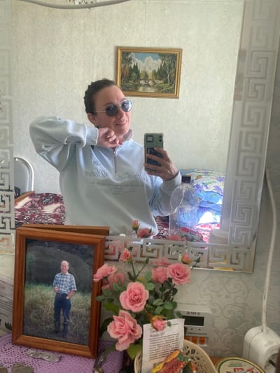 A mirror selfie taken in what appears to be a home interior.

**Person:** A person wearing round sunglasses and a light-colored zipped sweatshirt is taking the selfie. Their face is visible, and they are holding up a smartphone.

**Subjects/Objects:**
*   Mirror: A large mirror with a decorative patterned frame.
*   Wall: The wall behind the mirror is covered with patterned wallpaper.
*   Decorations: A framed picture with a portrait, artificial pink roses, and a basket are visible on a surface beside the mirror.
*   Other: There are also items such as a device (possibly an electronic control panel), and some paperwork.

**Actions:**
*   The person is holding up their phone to take a picture of themselves in the mirror.

**Environment:**
*   The setting appears to be inside a home with a traditionally decorated interior.

**Location:** This is not knowable based on the image. Узяр, Russia is mentioned in the instructions only.