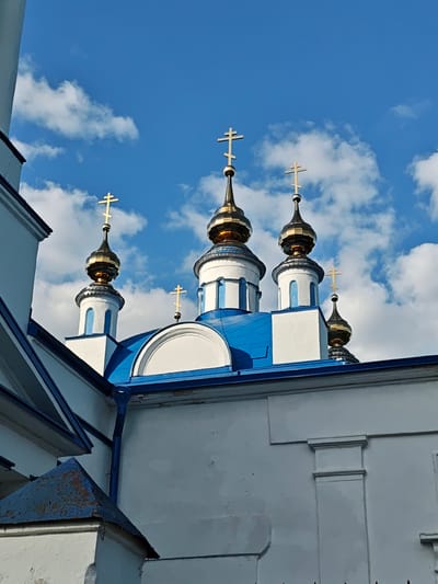 The image shows a white building with blue accents, topped with multiple gold-domed towers, each capped with a gold cross. The style suggests a Russian Orthodox church. The sky is bright blue with scattered white clouds. Based on the prompt, the location is Мамонтово, Russia.