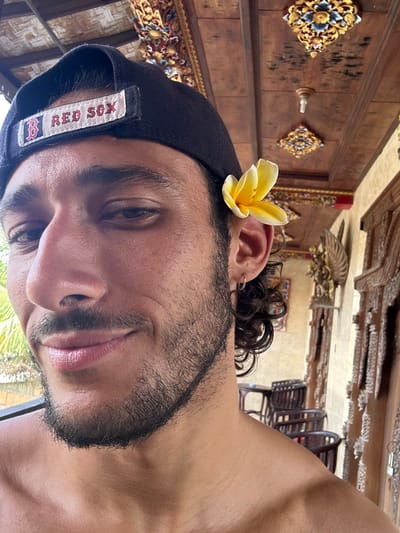 A close-up photo of a man with a beard wearing a black baseball cap with the words "RED SOX" on it. A yellow flower is tucked behind his ear. He gazes directly into the camera with a slight smile. The background consists of ornate wooden carvings on a wall and ceiling. The image is taken in a horizontal orientation.