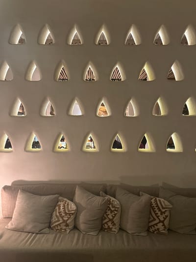 The image depicts an interior scene with a textured, light-colored wall featuring numerous triangular perforations. These perforations reveal glimpses of the adjacent space, suggesting a decorative or functional separation of areas. To the right of the wall is a sofa or seating arrangement with an assortment of pillows, some plain and others with geometric patterns. The lighting is soft and warm, highlighting the texture of the wall and the contours of the furniture. Given the location in Ramat Negev Regional Council, Israel, this may be part of a resort or residential setting that incorporates design elements appropriate for a desert environment.