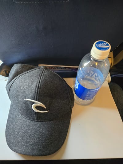 A dark gray baseball cap with a white embroidered logo rests on a white tray. To the right of the cap, a clear plastic bottle with a blue and white label and cap contains a light-colored liquid. The tray is placed on a dark blue surface, possibly a seat in a vehicle or aircraft. The background is dark and out of focus. The image appears to have been taken in a dimly lit environment. There is no indication of the location being Ramunia Dua, Indonesia.