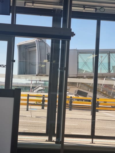 The image depicts a view through a window, likely at an airport gate. The exterior view shows a jet bridge connecting to an aircraft. The jet bridge has glass panels and yellow support structures. A vehicle with the "Swissport" logo is visible. The ground is paved and appears to be an airport tarmac. The sky is clear and blue. Based on the name of a company, the location might be near Saint-Louis, France (near the border with Switzerland and Germany).