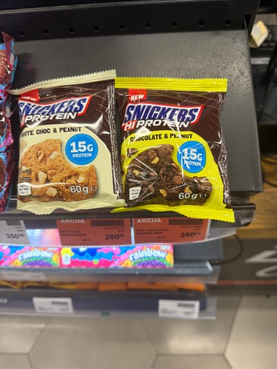 The image shows two different Snickers Protein bars displayed on a shelf. The top bar is chocolate and peanut flavored, and the bottom one is white chocolate and peanut flavored. Each bar is labeled with "15G Protein" and "60 g". In the background, there are price tags showing the prices of the Snickers bars and a "rainbow" item. The price for the chocolate and white chocolate Snickers bars is 280.00, marked as AKCIJA, which means "sale" in Serbian. The "rainbow" product is priced at 350.00. The location of the image is in a store somewhere in Stara Pazova Municipality, Serbia, as inferred from the Serbian language on the price tags.