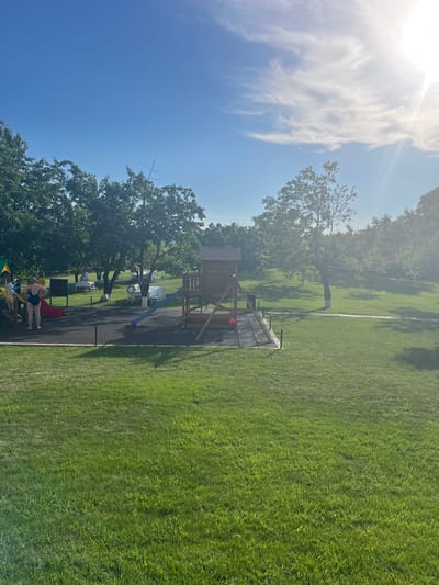 The image depicts an outdoor playground scene with a large grassy area and trees. The sun is high in the sky, indicating it is likely daytime, and the bright light suggests sunny weather with some wispy clouds. The playground structure is visible, featuring a wooden fort with a slide and an additional red slide. There are also picnic tables and chairs set up on the grass in the background, suggesting a place for recreation or an event. Two people are visible near the playground, one in a blue swimsuit and another in a pink swimsuit, appearing to be engaged with the play equipment. The overall setting seems to be a park or recreational area in Stăuceni, Moldova. The image has a bright and cheerful atmosphere due to the sunny weather. No text is visible in the image.