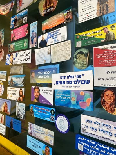 This image depicts a dense collection of memorial stickers and posters adhered to a dark, reflective surface, likely a wall or window, in the city of Kfar Vitkin, Israel. The scene is dominated by a grid-like arrangement of various-sized and shaped decals, primarily rectangular and circular, covered in photographs and Hebrew text.
Essential details include:
* **Subjects:** Numerous individuals, predominantly young adults, are depicted in color photographs across the various memorials. Many are smiling portraits; some males are shown in Israeli military uniforms.
* **Objects:** The primary objects are the stickers and posters themselves. They feature:
* Photographs of the deceased, typically front-facing portraits.
* Hebrew text, including names, dates (often birth and death years, such as "1988-2023," "2002-2024," or specific dates like "7.10.2023"), and memorial messages.
* Israeli flag motifs (blue and white colors, Star of David).
* QR codes, presumably linking to further information.
* One prominent circular sticker clearly reads "REMEMBER Bar Tomer 3.10.2023."
* A central white rectangular sticker contains a large Hebrew phrase which translates to "When will we all understand that we are brothers," or similar, indicating a collective message.
* **Actions/Interactions (implied):** The assembly of these memorials represents a public act of remembrance and mourning for individuals, many of whom appear to be victims of conflict or fallen soldiers. The presence of dates like "7.10.2023" specifically links some memorials to the October 7th attacks. The stickers are adhered flat against the surface, indicating a semi-permanent display.
* **Environment:** The background is a uniform dark, glossy surface, suggesting an indoor wall or a shop window. The lighting is consistent, illuminating the printed materials clearly. No other environmental elements like furniture or landscapes are distinctly visible beyond the immediate display surface.