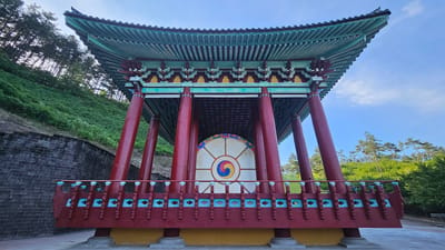 A traditional Korean pavilion with a large drum is pictured in Godang-ri, South Korea. The pavilion has a multi-tiered, green tiled roof with ornate red and teal trim. Red pillars support the roof and surround a large, circular drum with a white face and a colorful taegeuk symbol in the center. A decorative red railing with teal accents encloses the platform of the pavilion. The pavilion is situated on a hillside covered in lush green vegetation. A stone wall is visible to the left of the pavilion. Pine trees and other green foliage form the background, under a bright blue sky with scattered white clouds. No people are visible in the image.