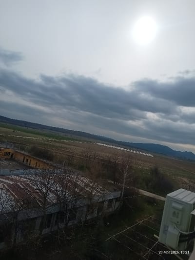 The image, dated March 20, 2026, at 15:21, presents an elevated, wide-angle view of an outdoor scene in Vedrare, Bulgaria. The sky occupies the upper two-thirds of the frame, characterized by extensive dark gray cloud cover, through which a brilliant, overexposed sun is visible in the upper right quadrant, casting significant glare.
In the immediate foreground, partially visible in the bottom right corner, is a white, rectangular telecommunication or utility box, mounted on a structure, with associated dark cabling. Below the horizon line, on the left side, a cluster of low-rise, single-story buildings with corrugated metal roofs appears dilapidated or abandoned. Bare-branched deciduous trees are scattered around these structures and in the immediate vicinity.
Extending from the area of the buildings towards the right and into the midground are vast expanses of brown, seemingly fallow or cultivated agricultural fields. Within these fields, a series of numerous white, elongated structures, consistent with greenhouses or polytunnels, are arranged in parallel rows. A faint dirt track or path is visible traversing the midground. The background features a range of distant hills or mountains, appearing dark green, situated beneath the cloudy sky. No human figures or discernible activities are present within the scene.