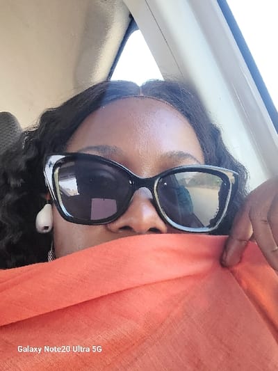 This image, captured in Rimin Ziem, Nigeria, displays a close-up of an adult individual inside a vehicle. The subject, possessing dark skin and dark, wavy hair, is visible from the forehead to the upper chest. They wear black cat-eye sunglasses with reflective lenses and a white wireless earbud in their left ear. An orange, woven fabric obscures the lower half of their face, from beneath the nose. The individual's right hand is visible, holding this fabric near the chin area. The background indicates a light-colored vehicle interior, featuring a headliner and A-pillar, with bright daylight visible through a window. A 'Galaxy Note20 Ultra 5G' watermark is present in the bottom left corner.