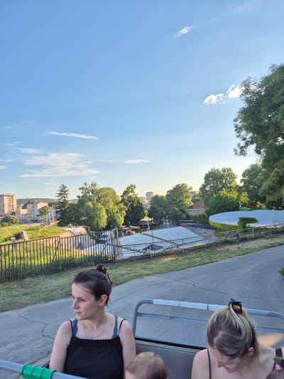 The image shows an outdoor seating area, possibly an amphitheater, with tiered white seats arranged in a semi-circle. A light blue sky with sparse white clouds is visible above. Trees and buildings, including a tall, light-colored building, are present in the background, suggesting an urban or semi-urban setting. In the foreground, two women are seated in a vehicle, possibly a tour bus or open-top vehicle. The woman on the left is wearing a black top and has her hair pulled back in a bun. The woman on the right has blonde hair with highlights and is wearing a dark-colored top. A child's head is visible between the two women. The overall scene appears to be a sunny day with good visibility. The presence of an outdoor seating area and the distant urban landscape suggests the location could be a park or a public venue within the city of Градска општина Црвени Крст, Serbia.