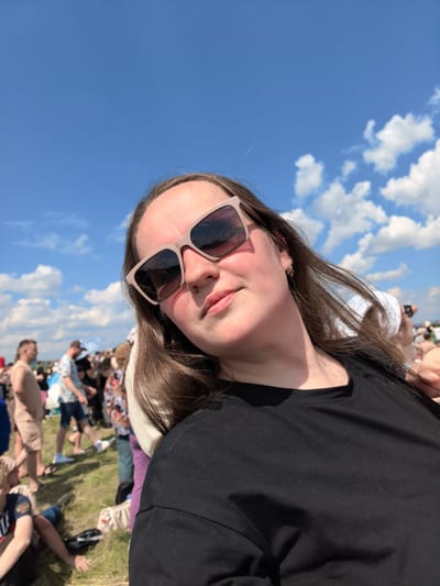 A woman wearing sunglasses and a black t-shirt is pictured in a selfie at a sunny outdoor event. The background shows a crowd of people and a blue sky with clouds. The location is identified as the city of Nestiukovo, Russia, suggesting a community gathering or festival. The woman is the primary subject, with the crowd and environment providing context.