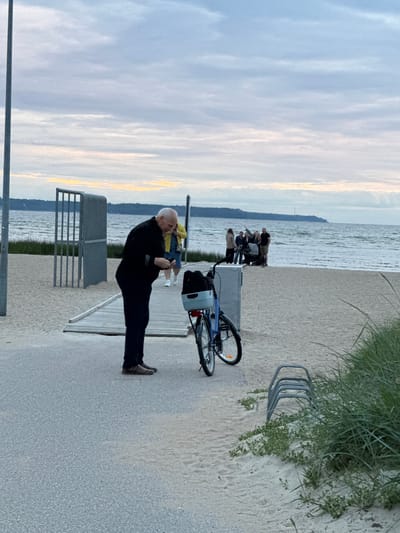 In the city of Tabasalu alevik, Estonia, an elderly man in a black sweater and dark pants is standing on a paved walkway next to a sandy beach. He is looking down at his phone. To his right, a blue bicycle with a basket is parked. In the background, a group of people are gathered near the water's edge. One person is pushing a stroller, and another is holding a dog on a leash. The sea is calm, and the sky is cloudy. The scene appears to be a public area with a walkway, beach, and some metal structures that may be part of a fence or barrier. There is also a bicycle rack visible in the foreground.