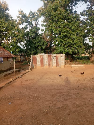 In Karsana 2, Nigeria, an outdoor scene depicts a modest building with a corrugated iron roof to the left, adjacent to a structure constructed of concrete blocks. This block structure features four metal doors, possibly serving as latrines or storage units. The ground is unpaved, composed of reddish-brown dirt with scattered pebbles. Three chickens are visible in the foreground, pecking at the ground. Lush green trees with dense foliage dominate the background, with a clear, light-colored sky above. The overall impression is one of a rural or semi-rural setting with basic infrastructure.