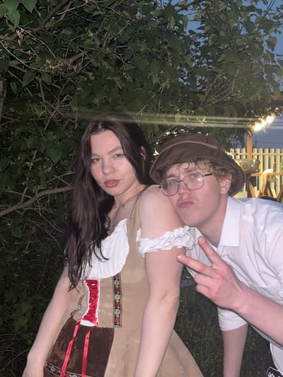 Two individuals are visible in the image. A male wearing a brown hat and glasses is leaning his head on the shoulder of a female. The female has long, dark hair. Both are wearing what appears to be traditional clothing, possibly related to Oktoberfest. They are positioned in an outdoor environment with a leafy tree visible in the background. The photograph seems to have been taken in Пальники, Russia. The man's hand is positioned, making a V shape.