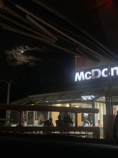 The image shows a night view of a McDonald's restaurant in Gelemenovo, Bulgaria. The restaurant sign is illuminated with the McDonald's logo and text. Several people are visible sitting at tables outside the restaurant. The moon is visible in the sky, partially obscured by clouds. The overall impression is of a typical evening scene at a fast-food establishment.
