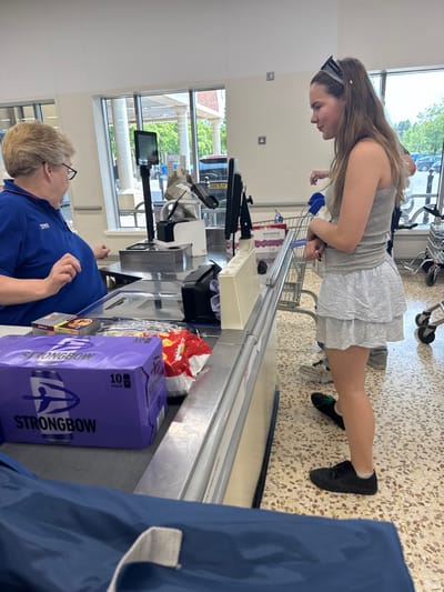 A young woman wearing a gray top and white ruffled skirt stands at a checkout counter with a shopping cart. An older woman in a blue Tesco shirt works at the till. A box of Strongbow cider and a bag of chips sit on the conveyor belt. The background features a window with a view of outdoor foliage and buildings. A blue shopping bag is partially visible in the foreground.