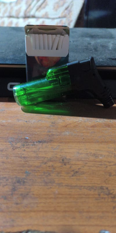 A pack of cigarettes and a green and black novelty lighter shaped like a gun are placed on a wooden surface. The cigarettes are white with a red and white pack. The lighter is transparent green with a black grip and trigger. The background is blurry and appears to be a table with a patterned cloth on it. The overall scene is dimly lit. The image does not contain any information that would allow for a determination of its location in the city of Есипово, Russia.